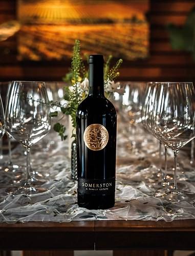 A bottle of Somerston red wine with the gold Astrolabe medallion, set on a table between empty wine glasses